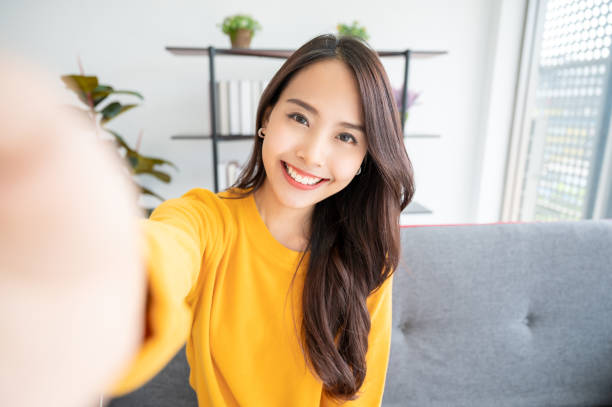 거실에 앉아 큰 미소와 예쁜 젊은 아시아 여성. 그녀는 흐린 배경에 빛 쾌활한 셀카를 찍는 재미를 가지고 - 셀카 뉴스 사진 이미지