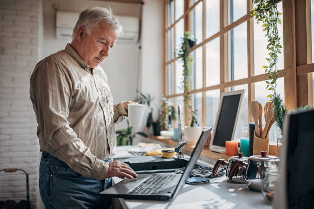 istock Senior man standing in the kitchen and working on the laptop while drinking his first coffee 1280080605