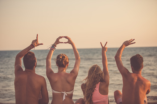 Rear view of four friends raising their arms to build the word LOVE with their hands. They sit facing the ocean during the sunset time at the beach.