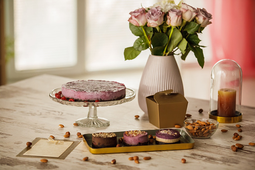 Shot of a table with a beautiful arrangement of different vegan cakes served on platters, love love letter, gift box and a bouquet of roses in vase with bunch of raw almonds scattered for decoration.