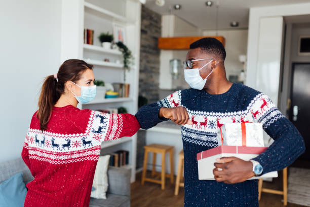 Young Caucasian woman welcoming her African American friend to her home Young Caucasian woman having her friend of African American ethnicity in her home for winter holidays, bringing gifts, wearing protective face mask, keeping social distancing and elbow bumping to greet each other social distancing photos stock pictures, royalty-free photos & images