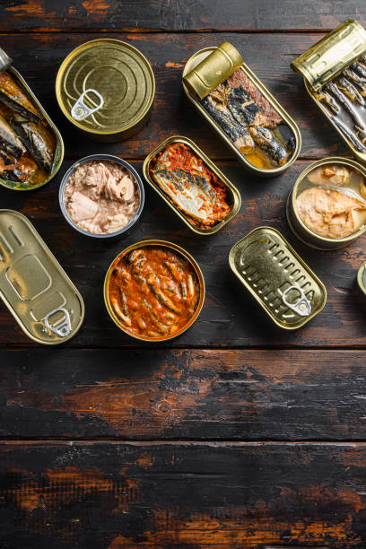 istock Tin cans for fish with different types of seafood, opened and closed cans with Saury, mackerel, sprats, sardines, pilchard, squid, tuna,  over  dark wood old table  flatlay top view  space for text 1280107480