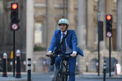 Front view of Caucasian corporate professional wearing full suit, helmet, and messenger bag approaching camera on bicycle while commuting to work.