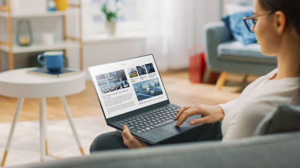 young woman at home utilizza il computer portatile per scorrere e leggere notizie sulle innovazioni tecnologiche. è seduta su un divano nel suo accogliente soggiorno. colpo sopra la spalla - pubblicazione foto e immagini stock