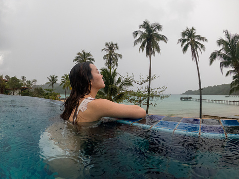 asian woman in luxury swimming pool looking away to the ocean in rainy day. travel bad weather concept.