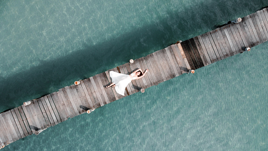 aerial view of asian woman in white dress lay down on wooden bridge over the transparent sea water near beach. copy space provided. travel concept. Thailand.