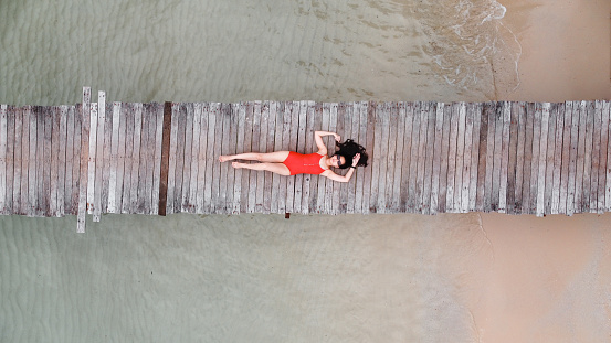 aerial view of asian woman in bikini lay down on wooden bridge over the transparent sea water near beach. copy space provided. travel concept. Thailand.