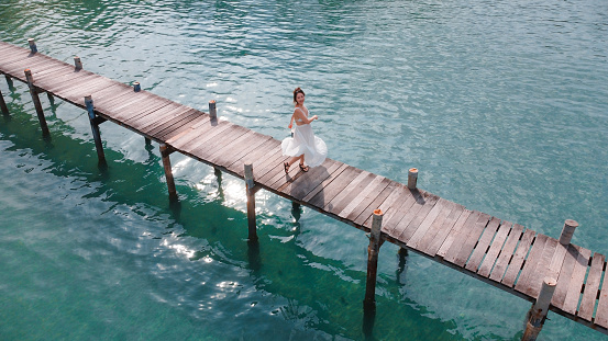 aerial view of asian woman in white dress walking on wooden bridge over the transparent sea water near beach. copy space provided. travel concept. Thailand.