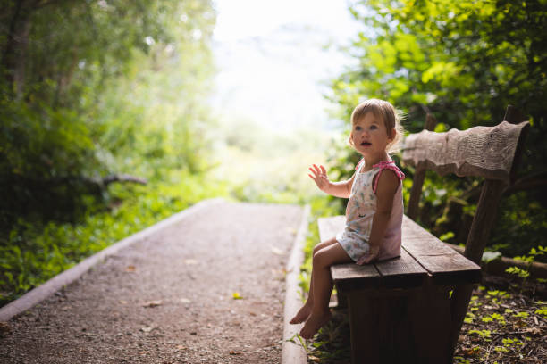 Female toddler barefoot with cute face sitting on wooden bench in nature park near path way with bright defocused background looking left, side view.