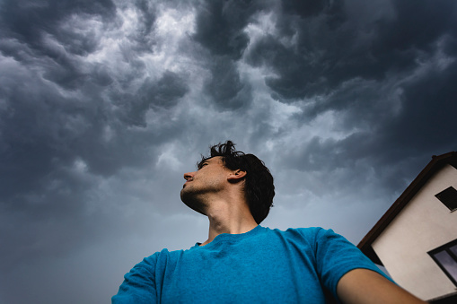 One young adult male selfie wide low angle of view of storm chaser looking up to dramatic sky over house in background for extreme weather phenomena.