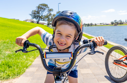 Cute cheerful boy with helmet on his new bike by the lake at the park. Lifestyle portrait of happy kid on his bicycle. Outdoor activities, vacations, happy childhood and back to normal concept.