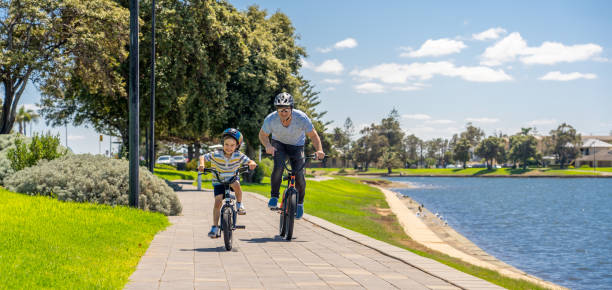 père et fils heureux conduisant leurs bicyclettes au bord du lac. père et fils s’amusant ensemble sur leurs vélos dans le parc. bonne famille, activités de plein air, l’enfance et le concept parental. - family bicycle cycling healthy lifestyle photos et images de collection