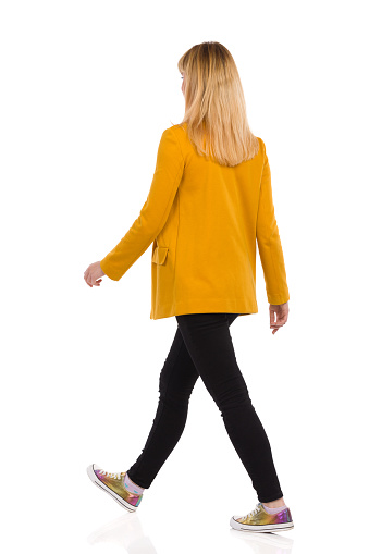 Rear view of walking young woman in yellow jacket, black jeans and sneakers. Full length studio shot isolated on white.