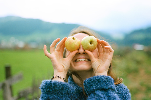 Young Caucasian woman placing apples on eyes while  walking in the village in Carpathian mountains
