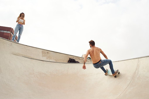 The man rides a skateboard in the bowl of a skatepark while his partner records him with the phone