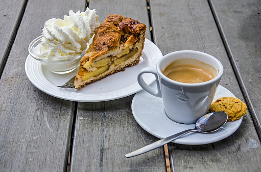 Wooden table with a cup of coffee with a biscuit as well as a saucer with apple pie and whipped cream