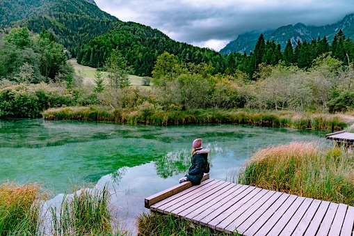 A young woman in a jacket is sitting on a wooden bridge and looking towards the lake. Crystal clear Lake Zelenci in cloudy morning. Slovenian mountains.