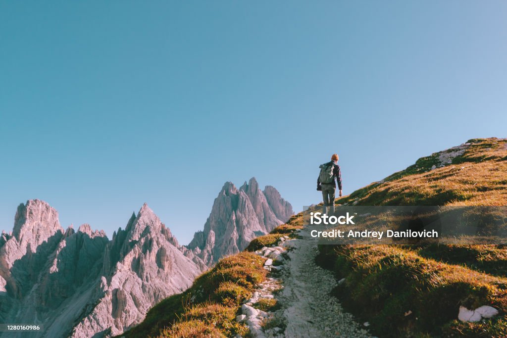 Man hiking alone on the edge of the rock and looking towards the horizon. Italian alps near the Tre Cime di Lavaredo. Young man with a backpack, wearing jeans, checkered shirt and yellow hat is hiking on the edge of rock. Rocky mountains and blue cloudless sky is on the background. Mountain Stock Photo