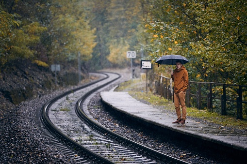 Pensive young man with umbrella waiting during rain at railroad station. Themes weather, autumn and loneliness.