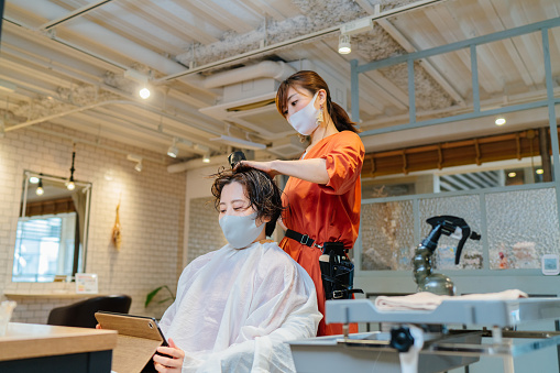 Hair dresser drying costumer's hair. Owner and customer wearing protective face mask for illness prevention
