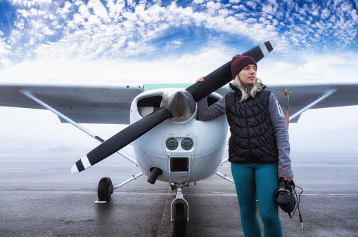Young Caucasian Female Student Pilot is standing in front of a Single Engine Airplane at the Airport. Taken during a foggy winter morning in Pitt Meadows, Vancouver, BC, Canada. Blue cloudy sky