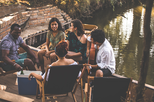 Full length shot of small group of diverse friends sitting near the river, hanging out, relaxing, playing guitar and enjoying their weekend getaway in nature together.