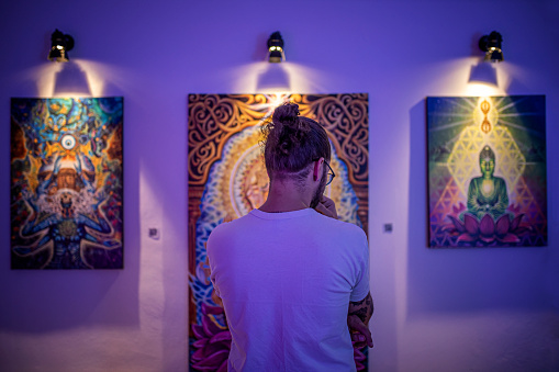 Rear view of young caucasian man in white shirt standing in an art gallery in front of colorful framed paintings displayed on a white wall