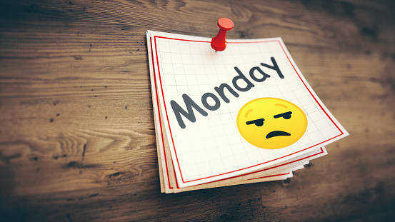 A stack of simple white note pages with the word Monday and an irritated face emoticon, on a wooden background. With copy space for additional composition.