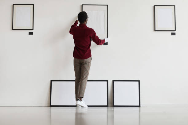 African Man Setting Up Frames in Museum Full length back view at African-American man hanging blank frames on wall while planning art gallery or exhibition, copy space hanging photos stock pictures, royalty-free photos & images