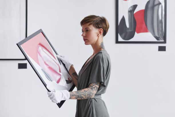 Creative Woman Holding Painting in Art Gallery Waist up portrait of tattooed creative woman holding painting while planning art gallery exhibition, copy space auction photos stock pictures, royalty-free photos & images
