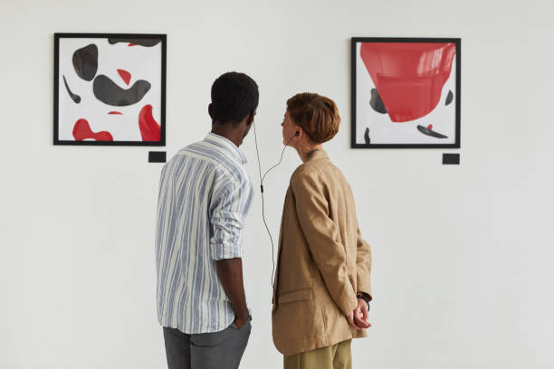 Couple Looking at Modern Art in Museum Graphic back view portrait of two young people looking at paintings and sharing audio guide while exploring modern art gallery exhibition, copy space museum stock pictures, royalty-free photos & images