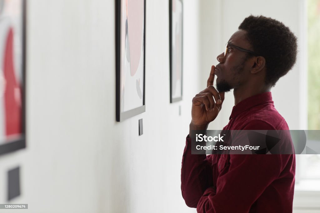 African-American Man Looking at Modern Art Side view portrait of young African-American man looking at paintings and thinking at art gallery or museum exhibition, copy space Museum Stock Photo