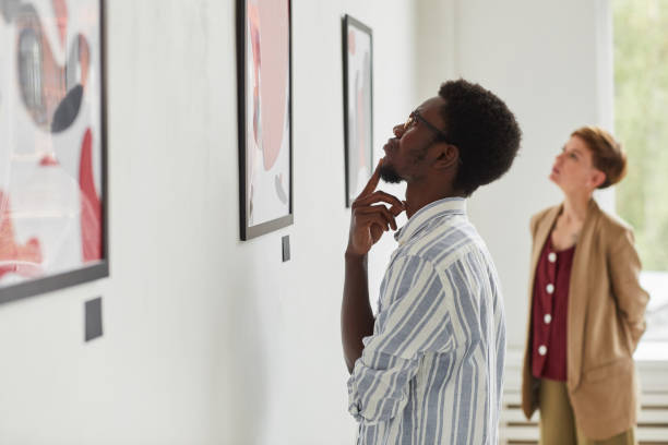 People Looking at Modern Art Side view portrait of young African-American man looking at paintings while exploring modern art gallery exhibition, copy space art museum stock pictures, royalty-free photos & images