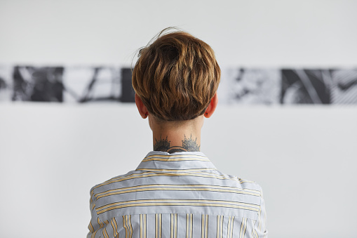 Graphic back view portrait of tattooed young woman looking at paintings at contemporary art exhibition, copy space