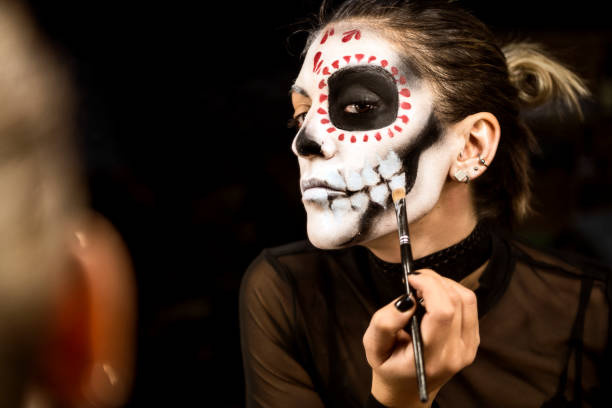 Young woman painting face for Halloween night party looking at mirror holding make up pencil Woman painting face for Halloween night party halloween face paint stock pictures, royalty-free photos & images
