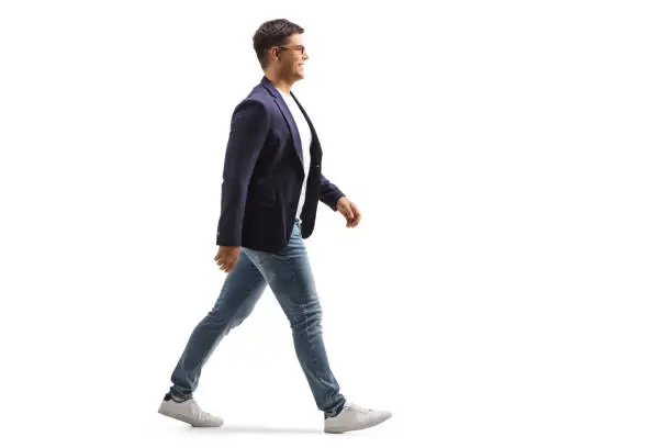 Full length profile shot of a smiling young man in jeans and suit walking isolated on white background