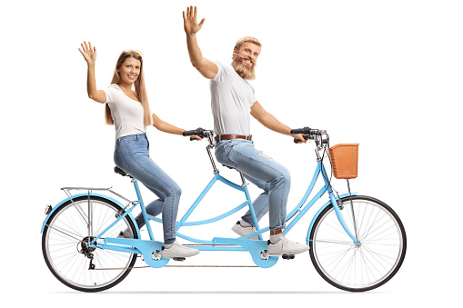 Young couple in matching casual outfits riding a tandem bicycle and waving at the camera isolated on white background