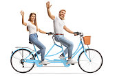Young couple in matching casual outfits riding a tandem bicycle and waving at the camera