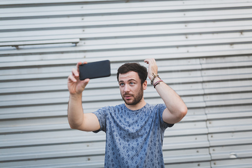 Handsome young man using a mobile phone for combing his hair while walking outdoors, taking a selfie.