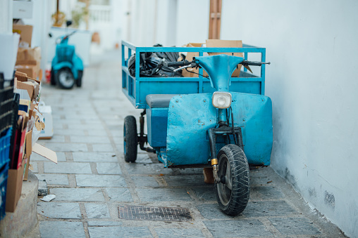 Blue three-wheeled old scooter with garbage bags and boxes near a white wall on a narrow street in the Lindos village on the Rhodes island, Greece.