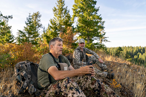 Two middle aged men wearing camouflage clothing rest on a mountain peak while hunting elk with a crossbow. It is a sunny Autumn morning