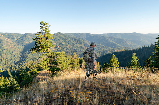 A middle aged man dressed in camouflage clothing and carrying a crossbow walks along a grassy ledge overlooking a forested mountain range in Washington state while hunting elk wild game.