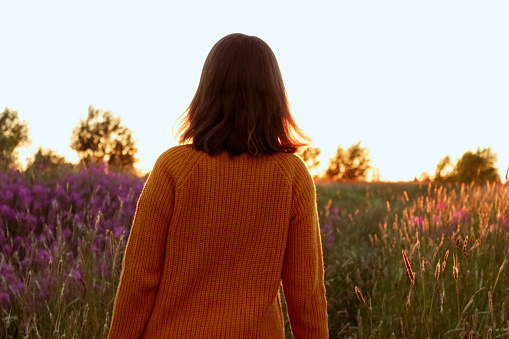 The girl stands with her back to the camera. Around her is a field, tall flowers and grass that glisten in the sunset.