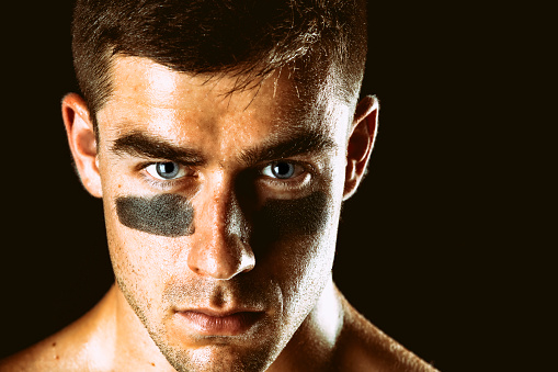 A close up of the face of an athlete wearing eye black and with sweat visible on the skin staring at the camera with a look of resolve and intensity.