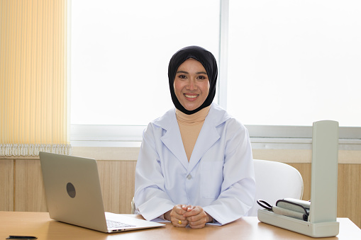 Female doctor wearing a hijab sits at a table waiting to examine a patient.