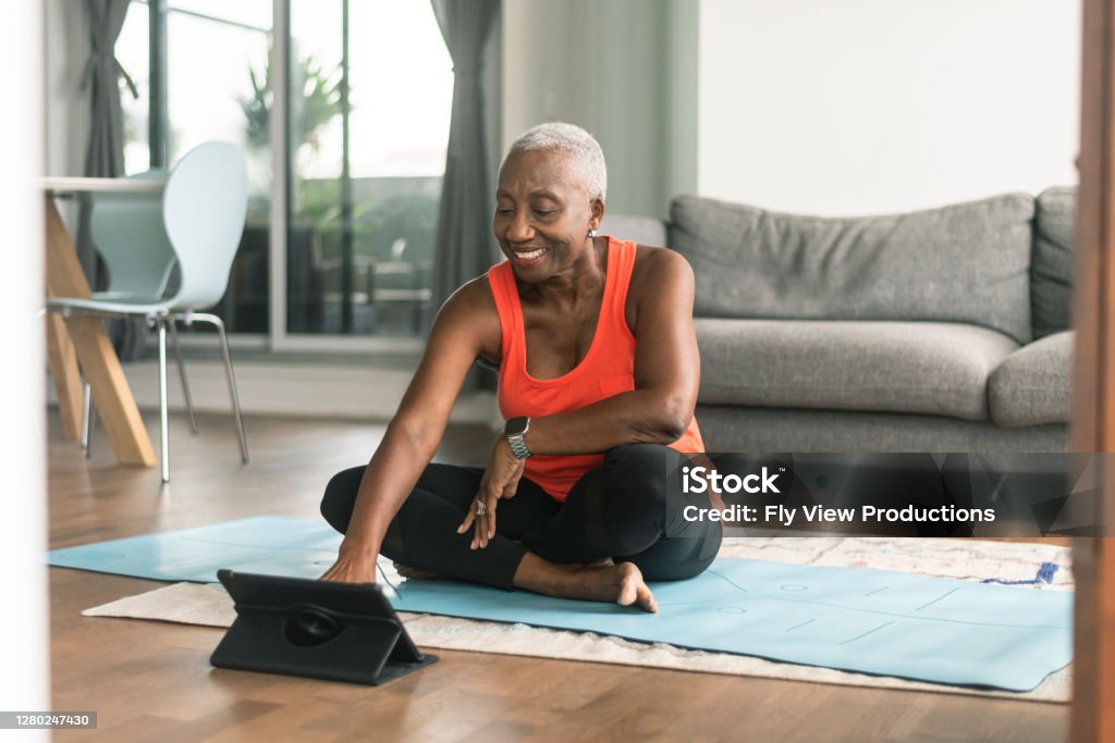 A black senior woman takes an online yoga class A beautiful senior woman takes an online yoga class. She is interacting with the teacher on her tablet and about to commence her class. She is wearing casual active wear and is taking the class in her lounge room. She is sitting on a blue yoga mat. Exercising Stock Photo