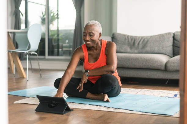 A black senior woman takes an online yoga class A beautiful senior woman takes an online yoga class. She is interacting with the teacher on her tablet and about to commence her class. She is wearing casual active wear and is taking the class in her lounge room. She is sitting on a blue yoga mat. exercise routine stock pictures, royalty-free photos & images