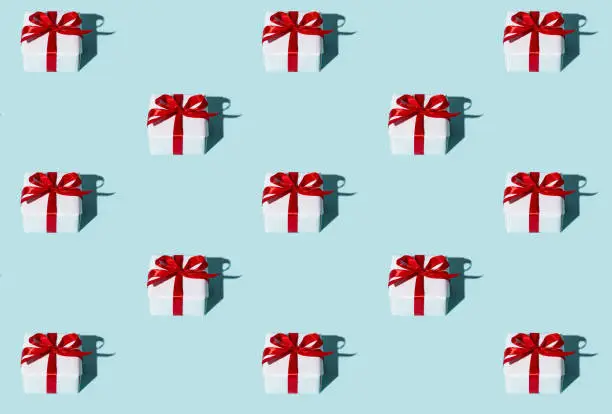 Gift pattern. Blue seamless background. Holiday surprise. Care package. Symmetrical arrangement of white boxes with red ribbon bows isolated on light.