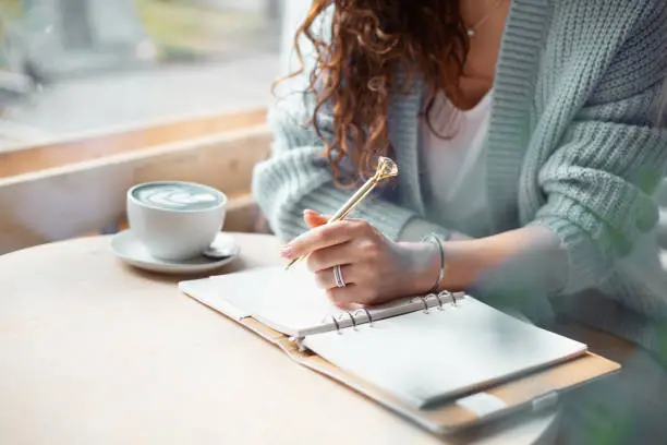 Photo of Young woman in blue warm sweater sitting near the big window of coffee shop and writing notes