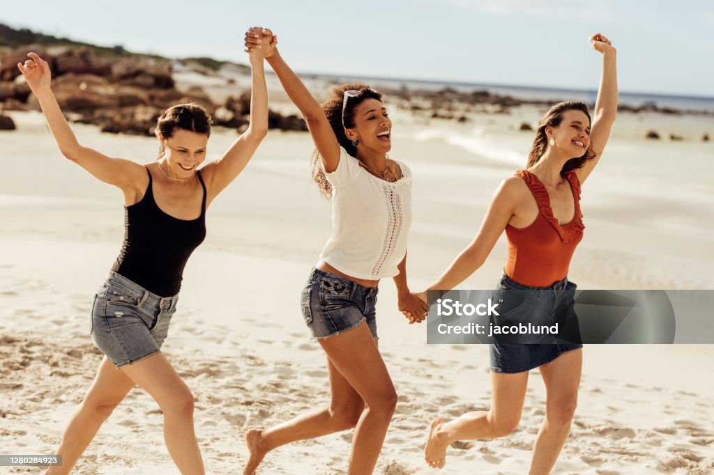 Friends enjoying beach vacation Happy women friends walking together on beach holding hands. Three young women enjoying and dancing together on a beach vacation. Beach Stock Photo